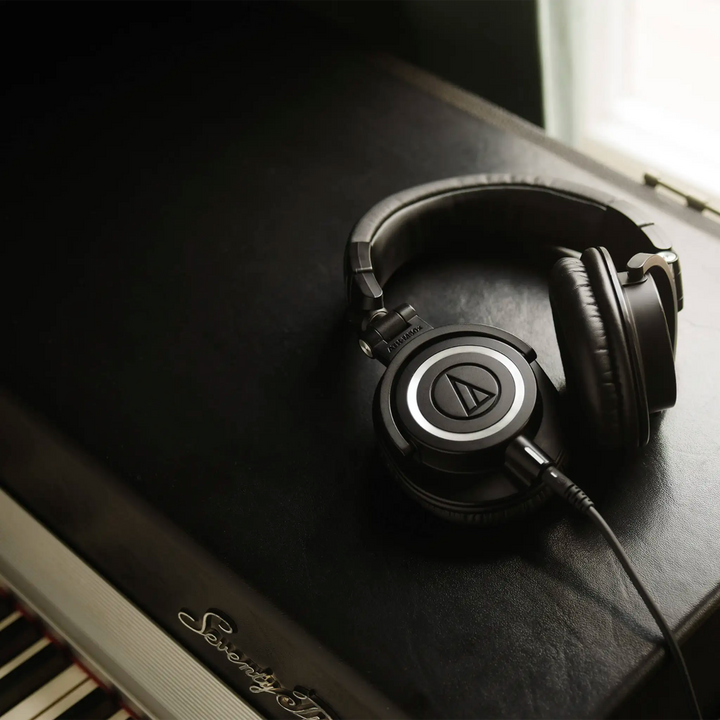 Audio-Technica ATH-M50x headphones lying right ear cup face up and left ear cup sitting up on leather padded piano top