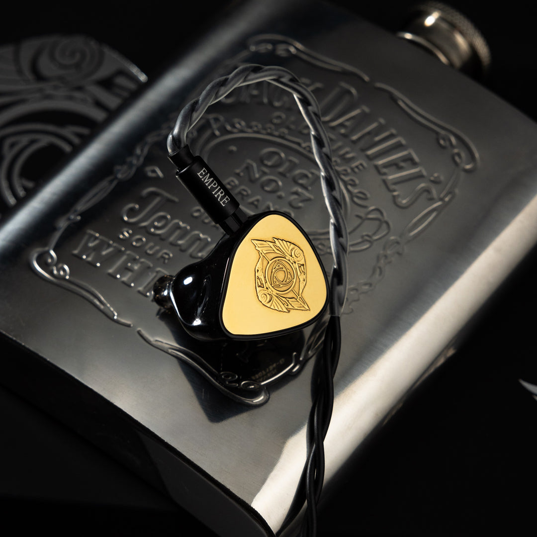 Empire Ears Raven gold launch edition single earphone with attached stock cable over Jack Daniels silver flask from ECT gallery