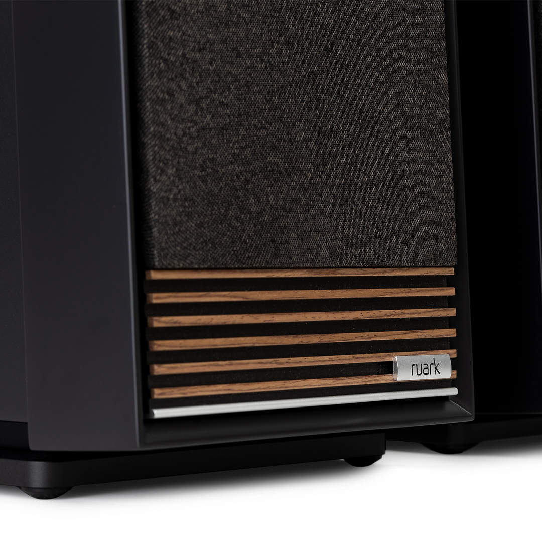 Close-up of a Ruark speaker cabinet showing the front grille with a dark gray woven fabric and horizontal wood slats, with a small Ruark logo badge.
