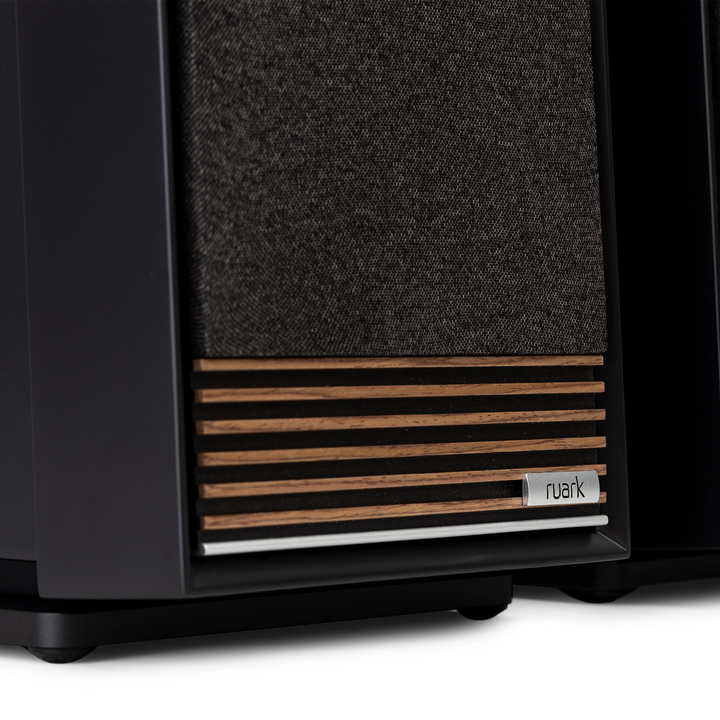 Close-up of a Ruark speaker cabinet showing the front grille with a dark gray woven fabric and horizontal wood slats, with a small Ruark logo badge.